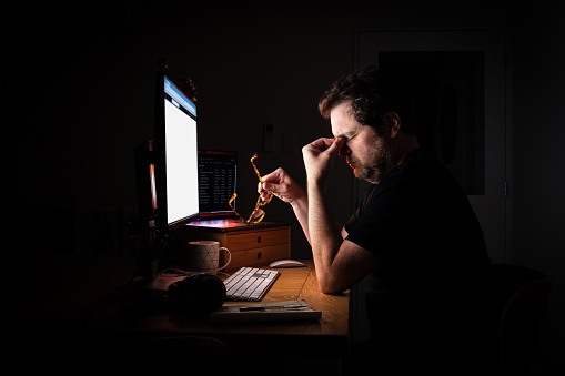 Tired and stressed office worker illuminated by the glow of their computer screen in a dark room at night while working late. Burn out.