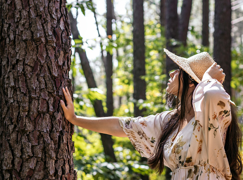 Young woman with a straw hat and dress walking through the forest, touching the trees, and feeling a connection with nature