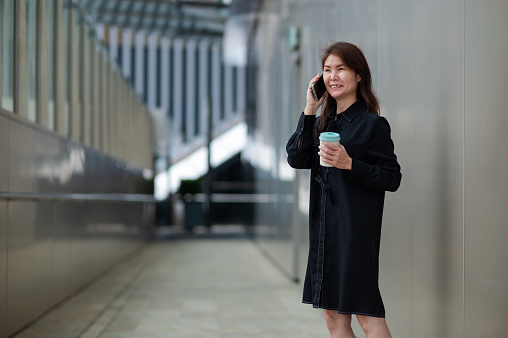 Asian female business executive in black suit using smart phone and holding a reusable coffee cup while standing outside the office building.