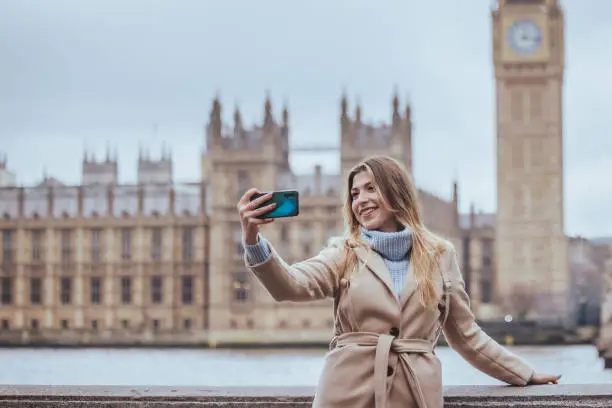 Candid portrait of a young cheerful blond female tourist in her 30s taking selfie with her smart mobile phone device while smiling wide with toothy smile visiting down town city of London, England. Selective focus with priority on the model with plenty of copy space on the background, which is defocused Thames river, Parliament building and Big Ben clock tower. Photo created during cold season outdoors and the model is with warm casual clothes in light cream and blue colours on a cloudy day with defused light  - creative stock photo