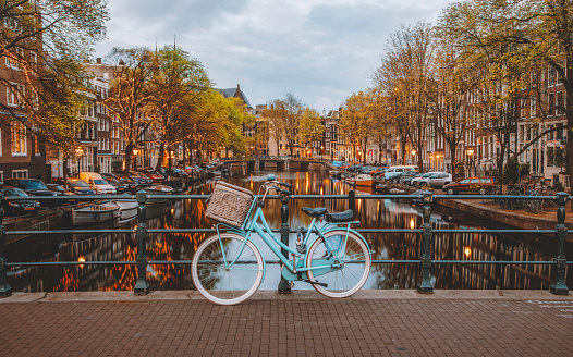 As early morning settles over Amsterdam, a solitary bicycle is parked on a canal bridge, silhouetted against the backdrop of illuminated city lights. The tranquil waters below reflect the warm glow of the street lamps, adding to the serene atmosphere of the scene. This captivating moment captures the timeless charm of Amsterdam's canal-lined streets, where the simple act of parking a bicycle becomes a picturesque vignette against the city's enchanting evening skyline in The Netherlands