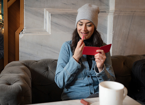 Woman applies lipstick as she looks at the pocket mirror for retouching in a cafe