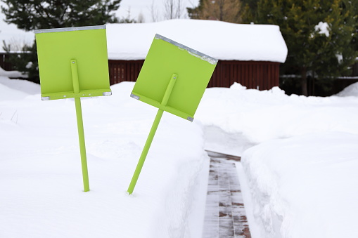 A two green wooden shovels for snow stand in a snowdrift near pathway in the courtyard of the country house on the background of winter landscape.