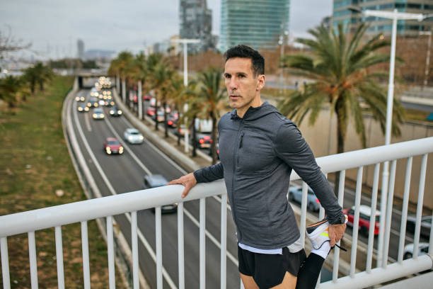 Three-quarter front view of sportsman in late 40s wearing workout attire, standing at railing of modern footbridge, preparing for afternoon exercise.