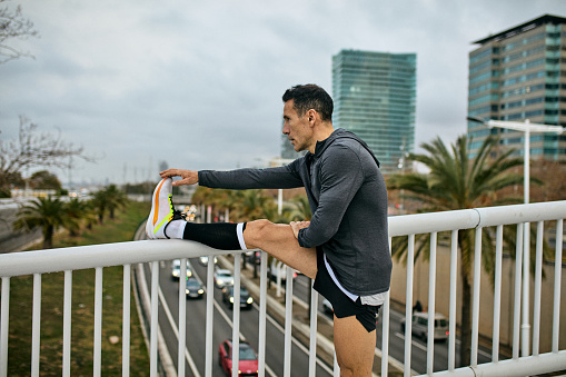 Side view of late 40s man wearing sports clothing, leg resting on footbridge railing, preparing for afternoon workout.