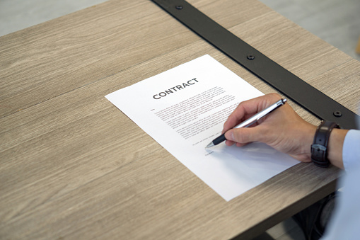 Closeup CONTRACT document on wooden table, man hand with wristwatch holding a pen signing on it.