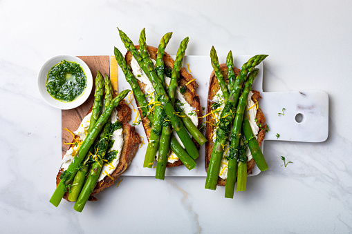Grilled toasts with sourdough bread with green asparagus, ricotta, lemon zest and herb oil on marble white board.