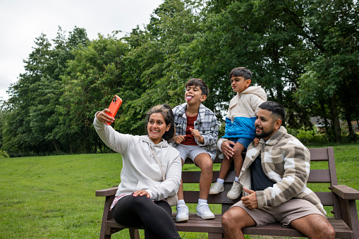 A mid adult couple and their sons enjoying a day outdoors in a public park in Ponteland, North East England. They are all taking a selfie together on a mobile phone and the children are pulling silly faces.