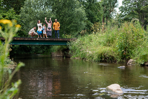 A multi-generation family enjoying a day outdoors in a public park in Ponteland, North East England. They are standing on a bridge above a river while bonding and throwing stones into it while looking excited with their arms in the air.