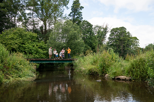 A multi-generation family enjoying a day outdoors in a public park in Ponteland, North East England. They are standing on a bridge above a river while bonding and throwing stones into it.