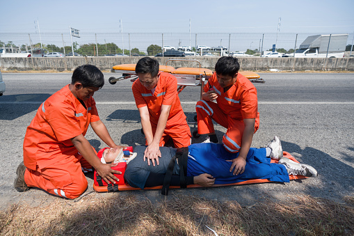 A man in head immobilizer tool lying on stretcher long spinal board with safety belt get CPR from rescue team. Urgent assistance during road accident.