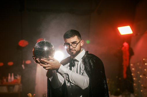Portrait of charming young man in a costume playing with a disco ball during a fun Halloween party. He is looking at camera and smiling joyfully.