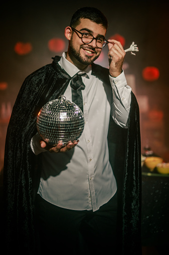 Portrait of charming young man in a cape for a costume, playing with a fake skeleton hand and a disco ball during a fun Halloween party. He is smiling at camera joyfully.