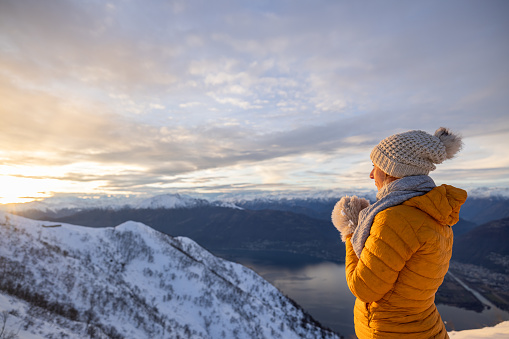Female hiker contemplates sunrise from mountain peak above lake in winter, on a high peak surrounded by snow