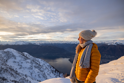 Female hiker contemplates sunrise from mountain peak above lake in winter, on a high peak surrounded by snow