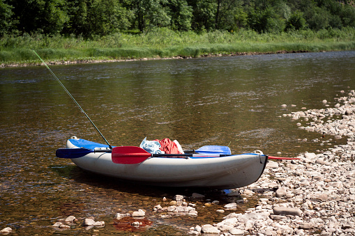 A watercraft known as a kayak, equipped with two paddles, is resting on the riverbank, by the shore of a flowing watercourse