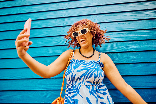Cheerful young woman with curly hair smiling while taking a selfie, wearing summer attire