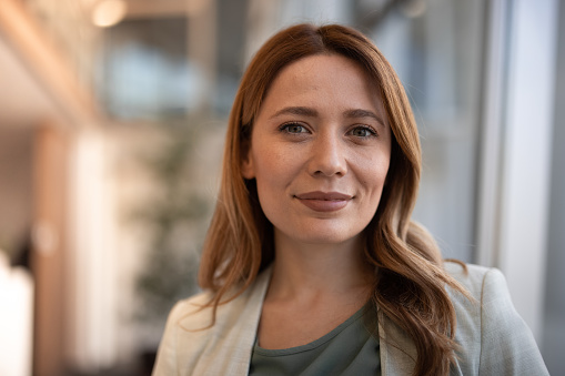 Closeup portrait of smiling beautiful middle-aged business woman wearing jacket and standing in light office hall with her arms crossed