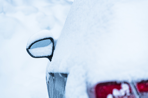 A cars side mirror is enveloped by a blanket of fresh snow, reflecting the chilly winter atmosphere in Sweden. The vehicle appears stationary, coated with snowflakes, emphasizing the cold, serene, and snowy conditions of the day.