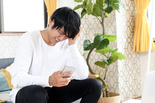 A young Asian man in casual clothes is looking at his smartphone indoors, holding his head in worry.