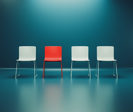 Four chairs stand next to each other in a row. The chairs are white, but one is red. Concept of choice, individuality and standing out from the crowd.