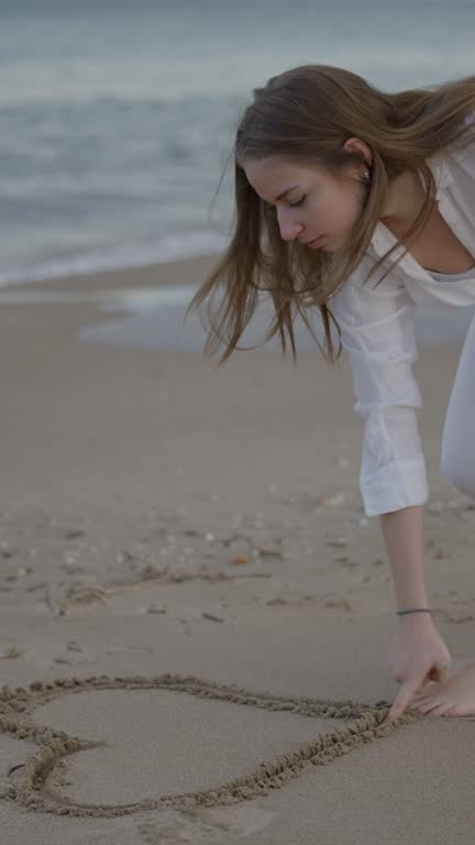 Cute young caucasian woman draws heart on the beach sand - slow motion shot