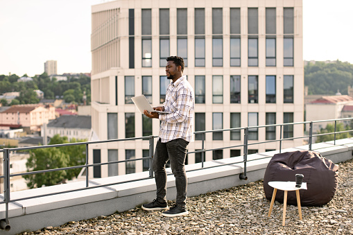 Full length view of African American businessman in casual wear standing on roof of modern office with laptop computer in hands during break.
