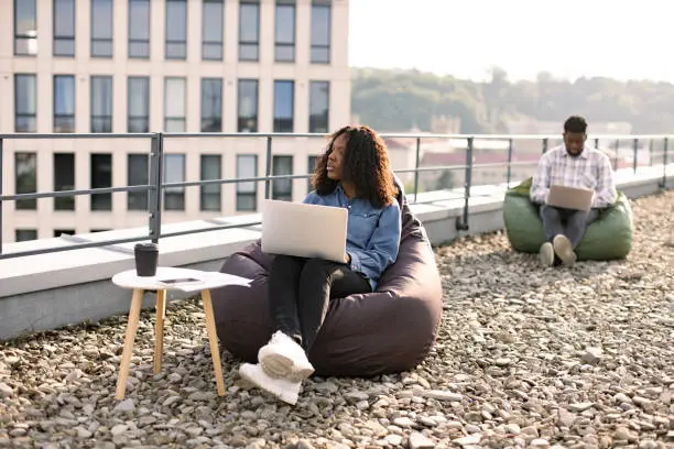 Successful business woman dressed in denim shirt and pants sitting at chair bag on roof top outdoors and typing on modern laptop, African American colleague works sitting in bag chair in background.