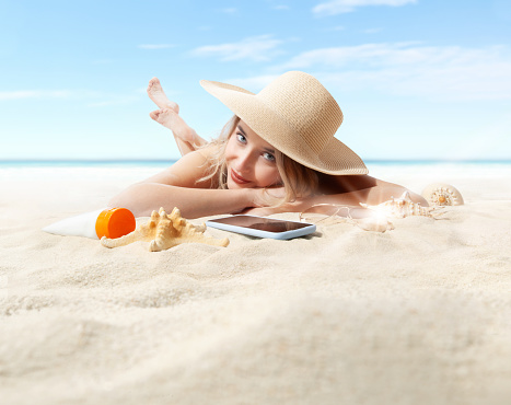 Smiling Woman lying on sand beach with smartphone, sunbathing with straw hat, concept of summer beach holiday and vacation travel. Sunscreen tube, sunglasses and seashell in foreground placed on sandy