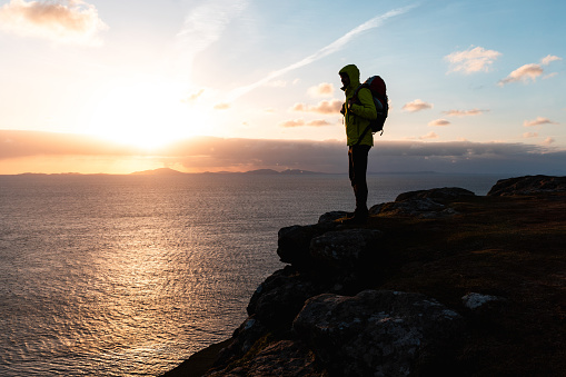 Explorer admiring scenic ocean sunset standing on cliff overlooking horizon - Silhouette of hiker carrying backpack enjoying solitude and reflection in the evening glow
