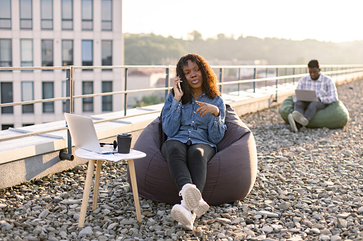 Carefree African American lady in casual wear making phone call while relaxing in bean bag on flat roof while male colleague working on blurred background.