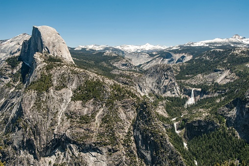 Side view of two mountain paths winding through a rugged and detailed landscape. The roads form a sharp visual contrast as they follow the natural shapes of the rocky ground. The asphalt's gray lines are prominent against the greenish rocky land. Parked by the side are cars that look tiny from afar.