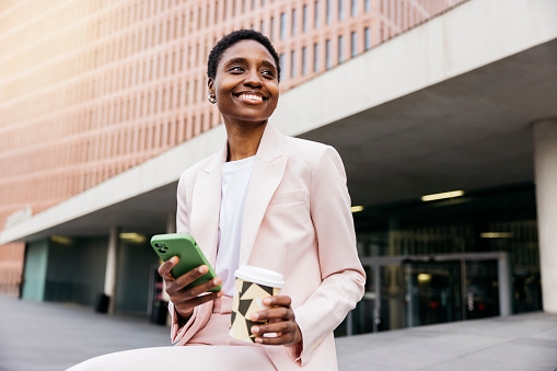 Young beautiful smiling businesswoman holding reusable mug and using a green phone during a coffee break in street in city.