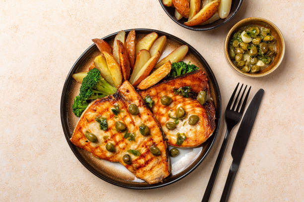 istock Grilled swordfish with garlic, lemon and capers butter sauce, with baked potatoes and broccoli. Top view, beige table, copy space. 2042720607