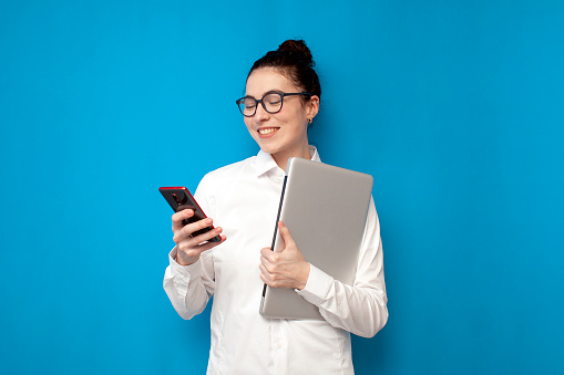 successful business woman in white shirt and glasses holds laptop and uses smartphone on blue background, girl manager financier is typing on phone on colored background, portrait of office worker