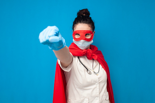 cheerful young girl doctor in uniform in superman costume on blue background, female nurse superhero shows forward and strength, healthcare concept