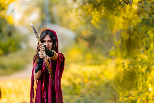 Portrait of a young magical woman preparing to fire an arrow with her bow in the forest.