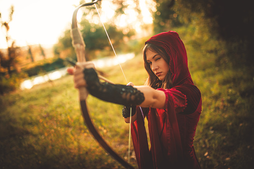 Young magical woman preparing to fire an arrow with her bow in the forest.