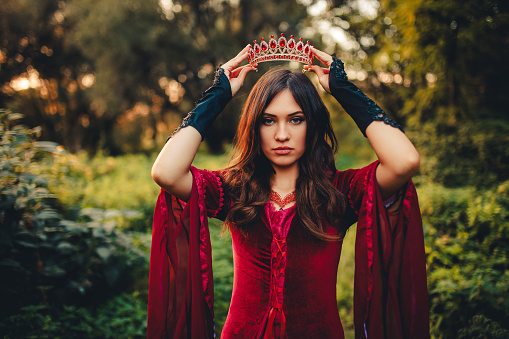 Portrait of a young woman dressed like an evil queen and putting a crown on her head in the forest.