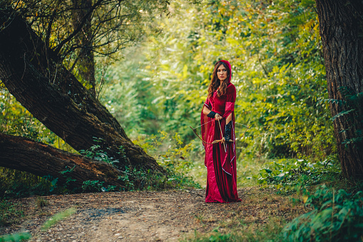Young magical woman preparing to fire an arrow with her bow in the forest.