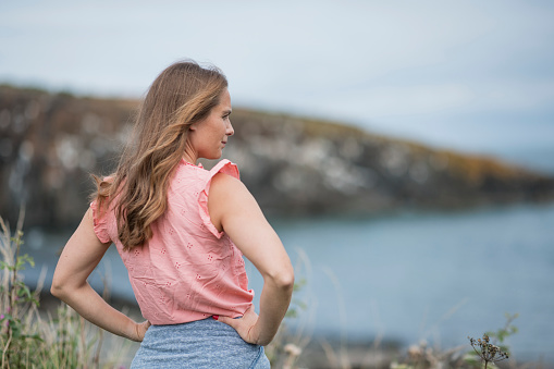 An over the shoulder view of a woman who is stood looking out to the sea in Craster in the North East of England. She has her hands on her hips and taking a moment to herself and appreciating the coastal views. She is dressed in summer clothing.