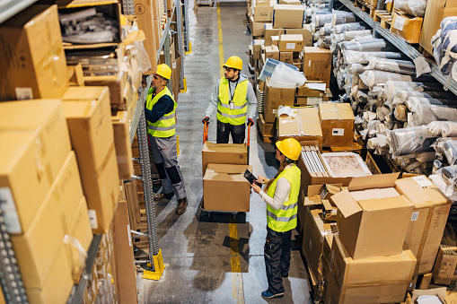 Two workers in a warehouse place cardboard boxes on a hand truck and prepare a shipment, as instructed by a female manager using a digital tablet