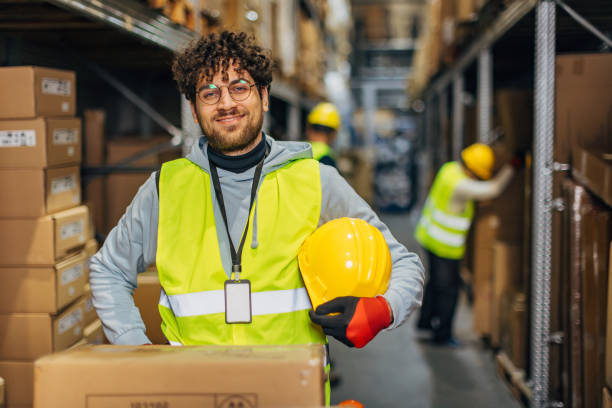istock Smiling warehouse worker holds safety helmet in hands while standing in warehouse and looking directly at camera 2042862279