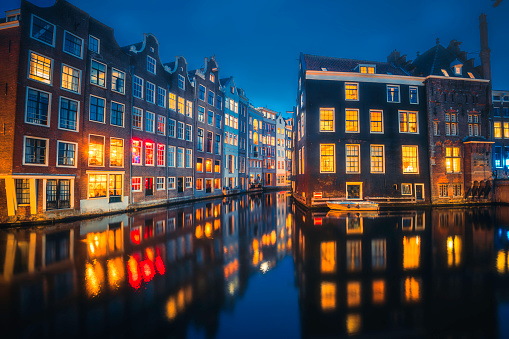 As night descends upon Amsterdam, the iconic canals shimmer with the warm glow of illuminated merchant's houses lining Oudezijds Voorburgwal and Zeedijk. This enchanting scene captures the timeless beauty and magical atmosphere of Amsterdam's historic waterfront during the evening hours in Holland