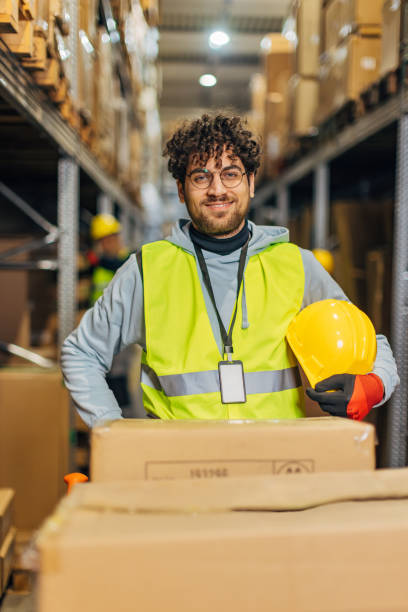 istock Smiling worker in uniform, wearing protective gloves and holding safety helmet in hands while standing in warehouse and looking at camera 2042876847