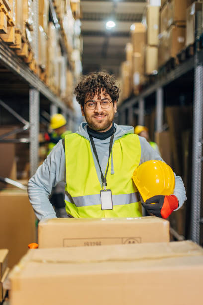 istock Worker in uniform, wearing protective gloves and holding safety helmet in hands while standing in warehouse and looking at camera 2042876850