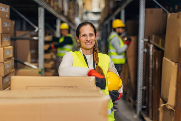 istock Female worker in uniform, wearing protective gloves and holding safety helmet in hands while standing in warehouse and looking at camera 2042877921