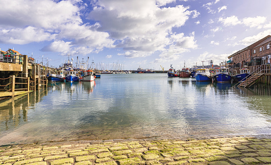 Scarborough, UK.  February 19, 2024.  Fishing boats are moored on two sides of a harbour with a marina in the background. The sea laps over a cobbled slipway and a sky with cloud is above.