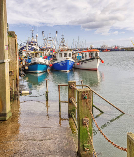 Scarborough, UK.  February 19, 2024.  Three fishing boats moored in harbour. A path with wooden posts and chains leads down to the water’s edge.