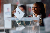 Happy black woman putting ballot into voting box at polling place during US elections.
